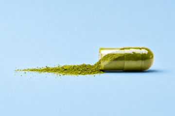 Geöffnete Kapsel mit grünem Matcha-Pulver auf hellblauem Hintergrund, daneben verstreutes Pulver